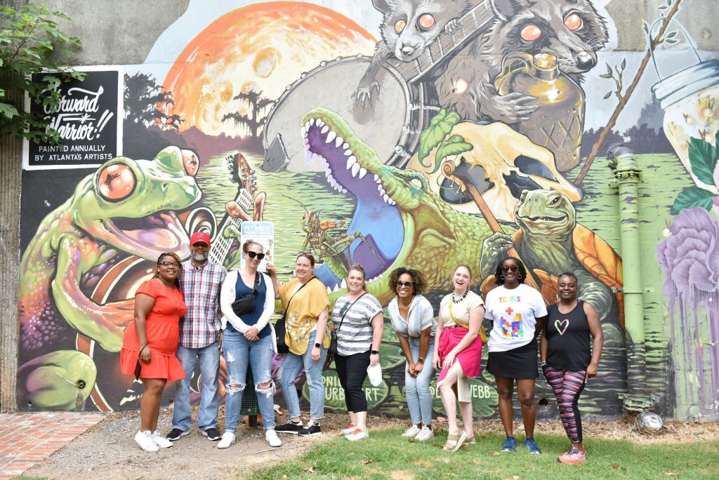 A diverse group of eight people stands in front of a vibrant mural featuring a whimsical swamp scene with animals playing instruments, smiling at the camera in Atlanta, Georgia.