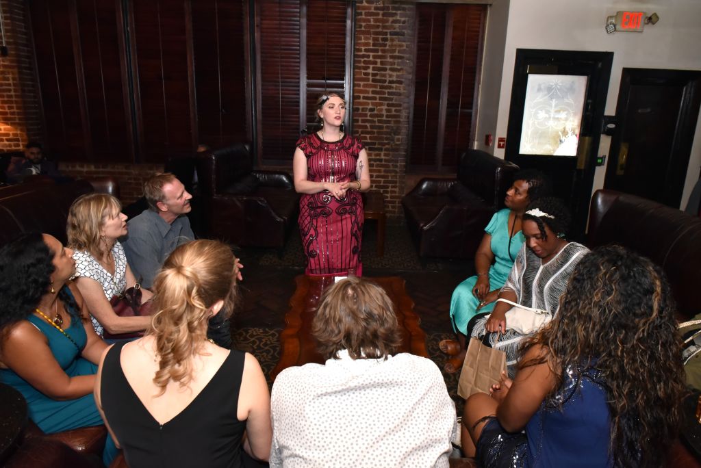 A diverse group of people is gathered in a cozy room with a brick wall and leather couches, attentively listening to a woman in a red dress who stands at the center, suggesting an intimate discussion or storytelling session in Atlanta, Georgia.