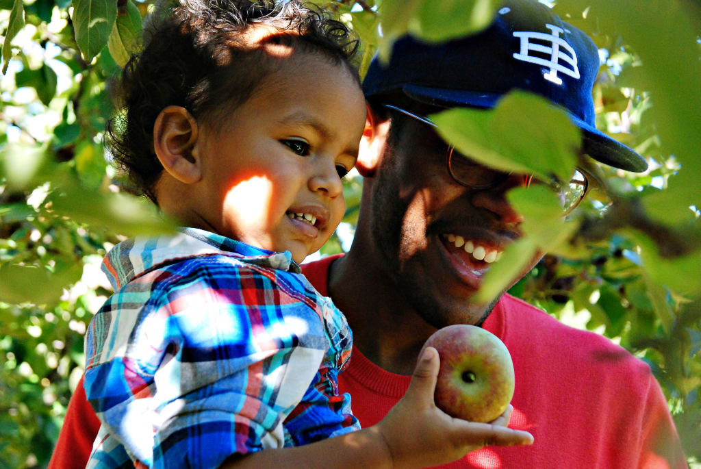 A smiling man wearing a baseball cap holds a child in an apple orchard in Atlanta, Georgia.
