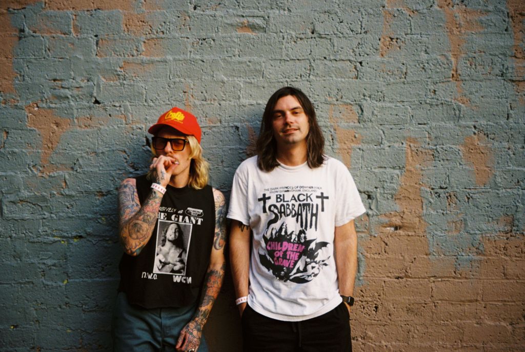 Two individuals stand against a weathered brick wall, one wearing a black tank top with a graphic and an orange cap, the other in a Black Sabbath t-shirt in Atlanta, Georgia.