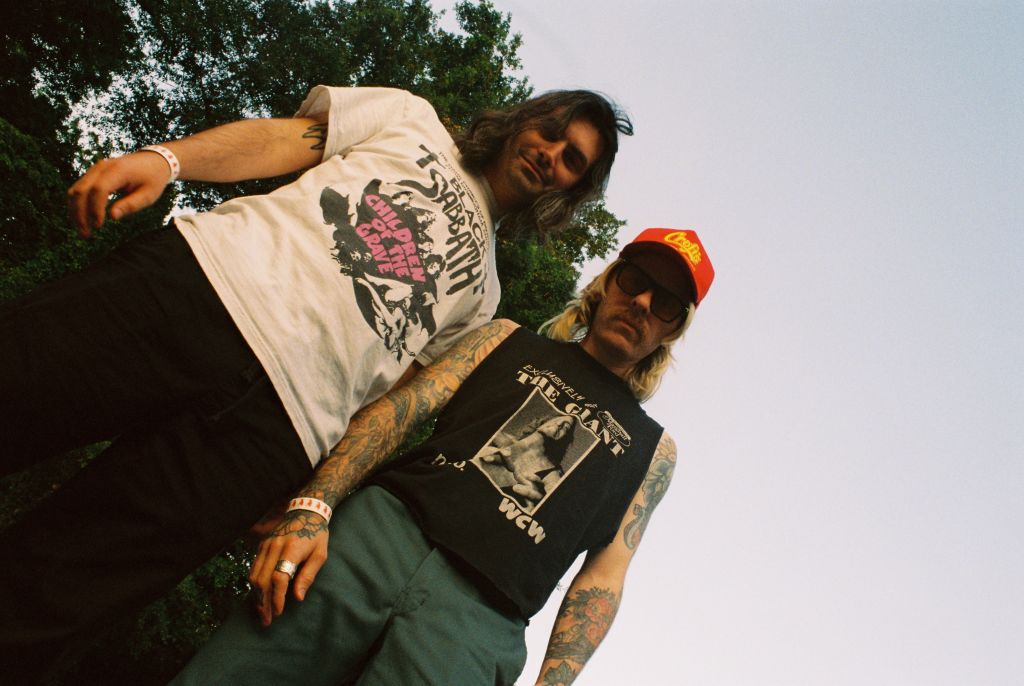 Ultimate Guide to Atlanta's Indie Music Scene 6 Two men with long hair and tattoos stand outdoors, one wearing a graphic t-shirt and the other a tank top, both sporting wristbands and rings in Atlanta, Georgia.