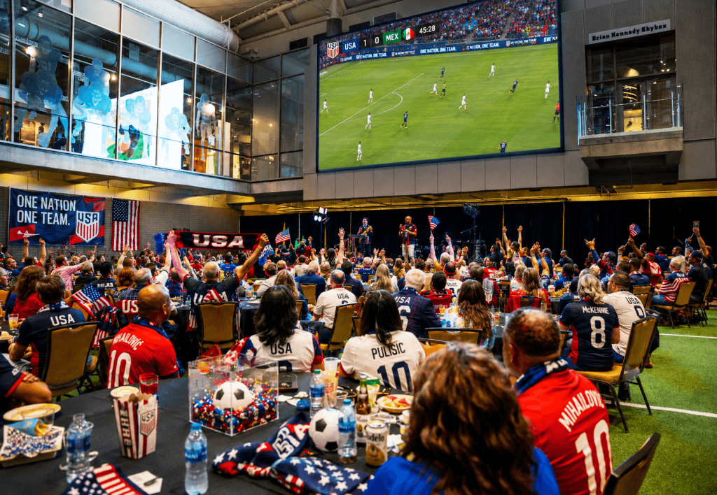 A large crowd of USA soccer fans, dressed in red, white, and blue, cheer and wave flags while watching a match on a giant screen in a convention center in Atlanta, Georgia.