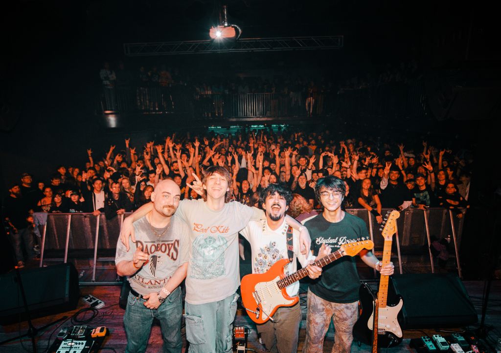 A group of five friends, four men and one woman, celebrate on stage with guitars, surrounded by an enthusiastic crowd at a concert in Atlanta, Georgia.