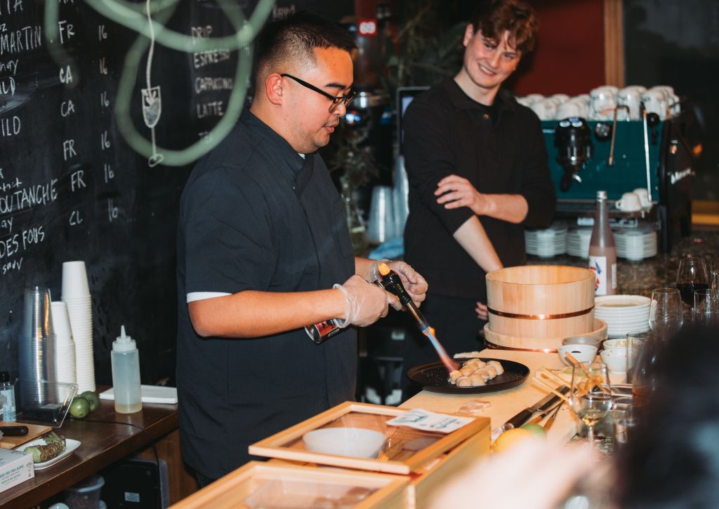 A chef in a black uniform is using a blowtorch to sear food on a plate, while another person stands nearby with arms crossed in Atlanta, Georgia.