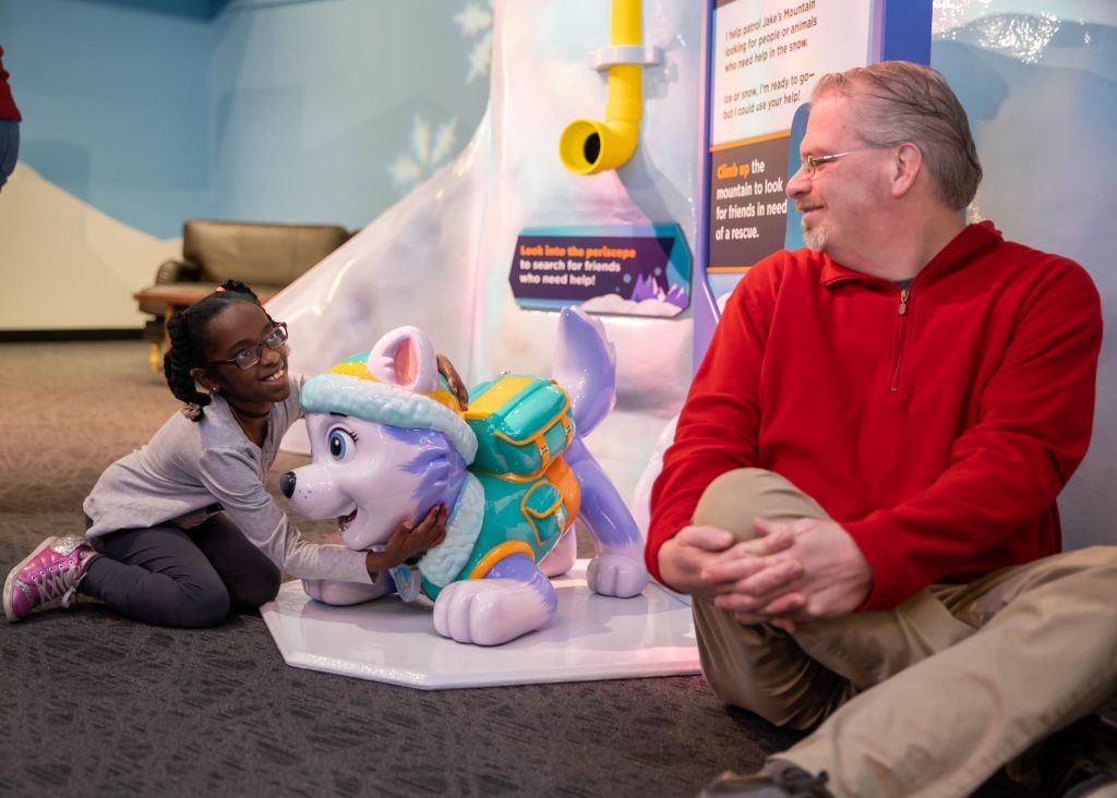 A man smiling at a little girl while she hugs one of the dogs from the PAW Patrol exhibit at Children's Museum of Atlanta. 