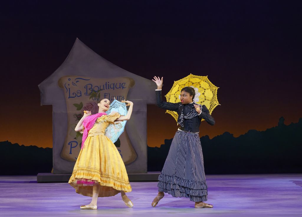 Two dancers in vibrant period costumes perform a lively dance on stage, one holding a blue fan and the other a yellow parasol in Atlanta, Georgia.