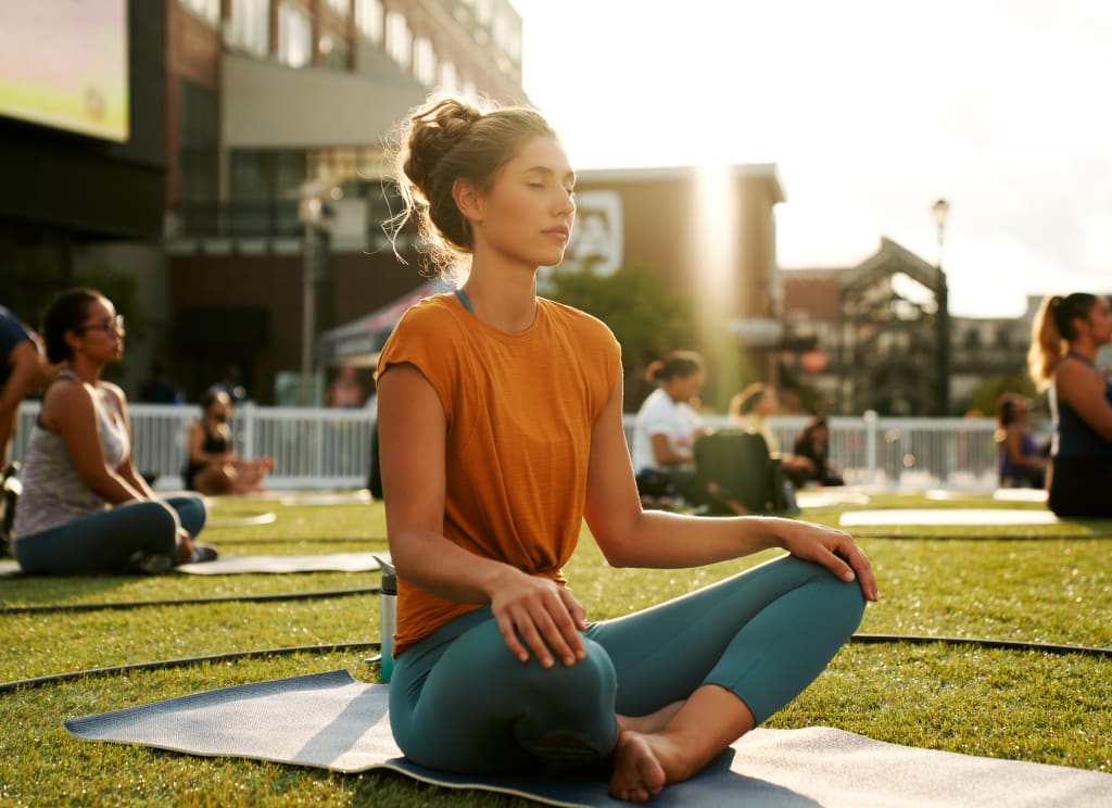 Woman meditating in lotus pose on a yoga mat in a grassy outdoor area with other people in Atlanta, Georgia.