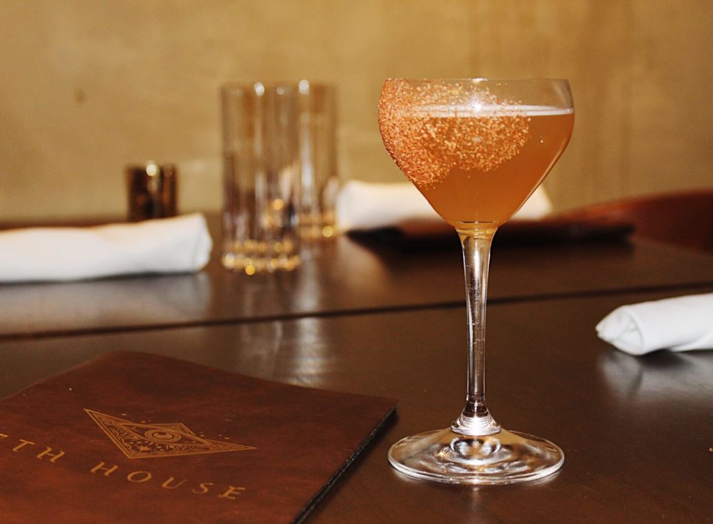 Must-Try Summer Cocktails in Atlanta 1 A martini glass filled with a golden-hued cocktail, rimmed with a reddish-brown sugar, sits on a dark wooden table next to a leather-bound menu with the words 'House Menu' embossed on it in Atlanta, Georgia.