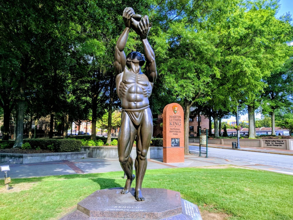 A muscular bronze statue of a man holding a sphere aloft stands on a pedestal in a park in Atlanta, Georgia.