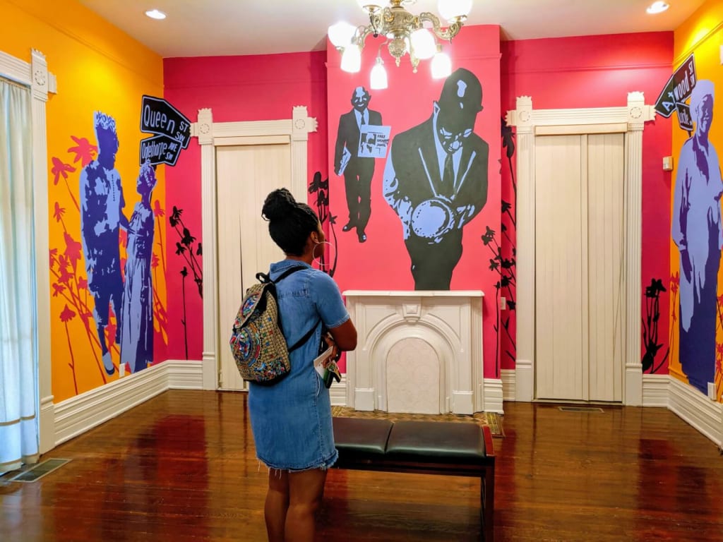 Woman with colorful backpack stands in vibrant room with murals and chandelier in Atlanta, Georgia.