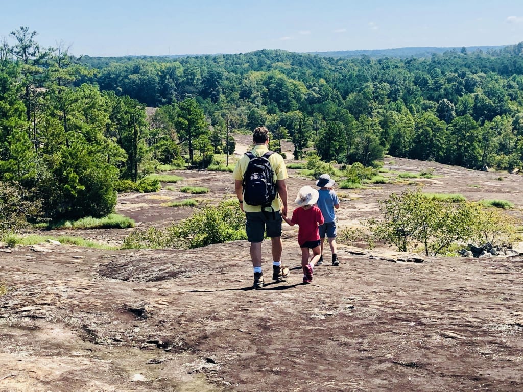 A man and two children hike on a rocky trail through a lush, green forest landscape in Atlanta, Georgia.