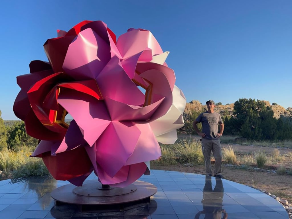 A man stands beside a large, colorful, abstract flower sculpture in a desert landscape in Atlanta, Georgia.