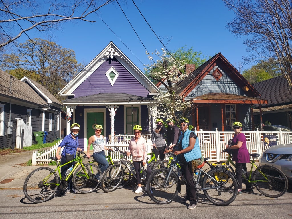 A group of cyclists wearing helmets and masks pose with their bikes in front of colorful houses in Atlanta, Georgia.