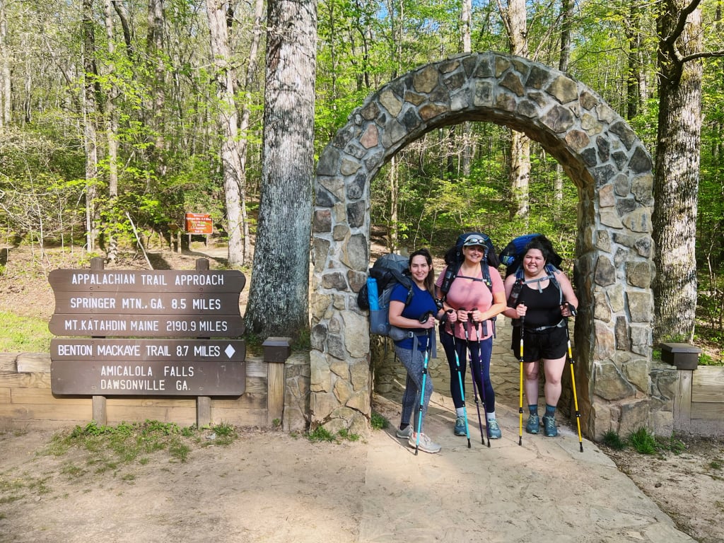 Three women smiling under the arch at the beginning of the Appalachian Trail Approach.