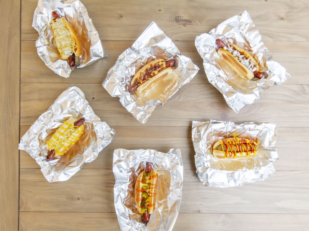 Six hot dogs with various toppings are wrapped in aluminum foil on a wooden table in Atlanta, Georgia.