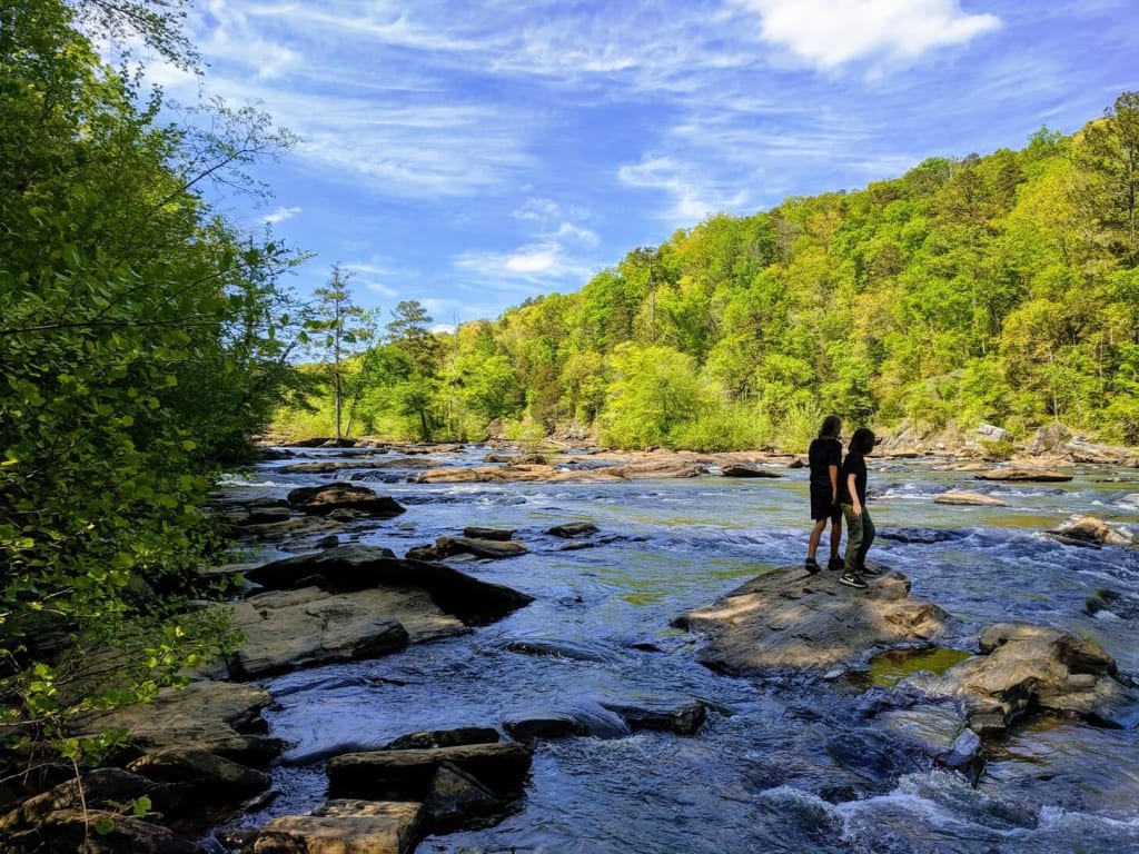 Two people stand on rocks in a river, surrounded by lush green trees and a clear blue sky in Atlanta, Georgia.
