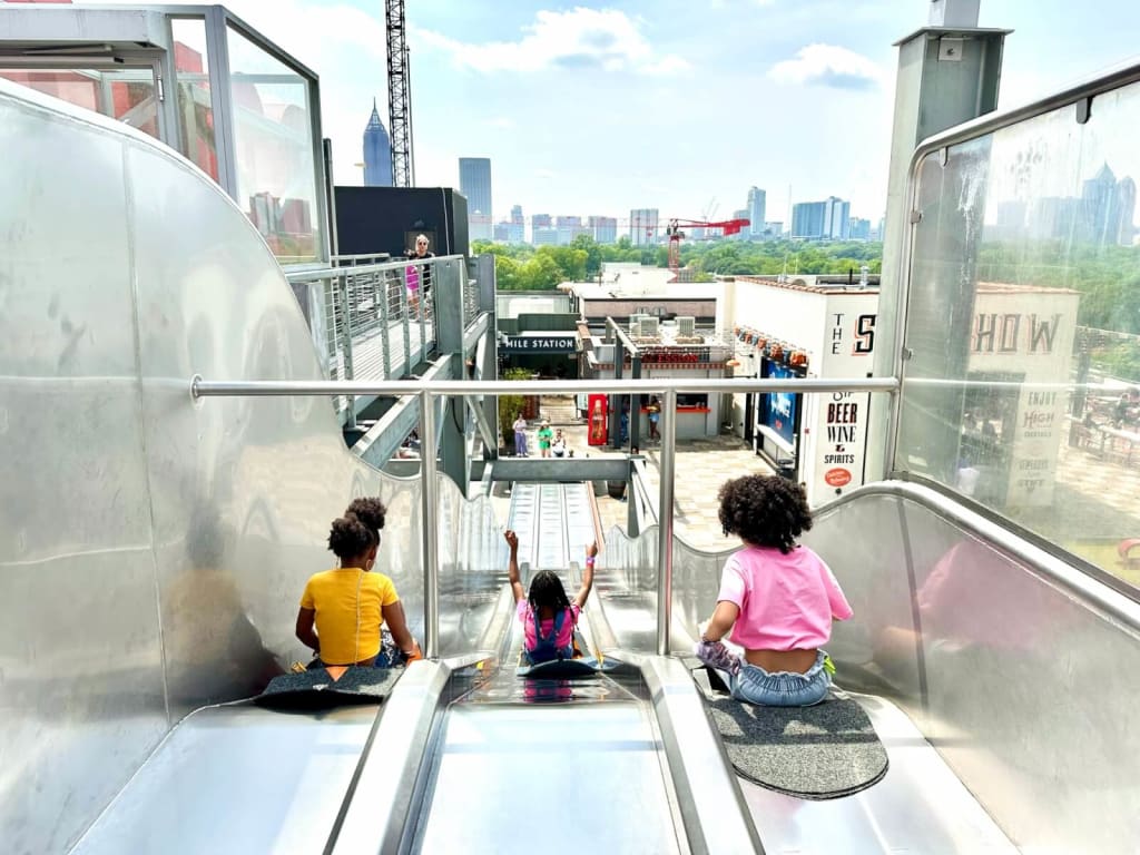 Three children slide down a long, shiny metal slide at an amusement park in Atlanta, Georgia.