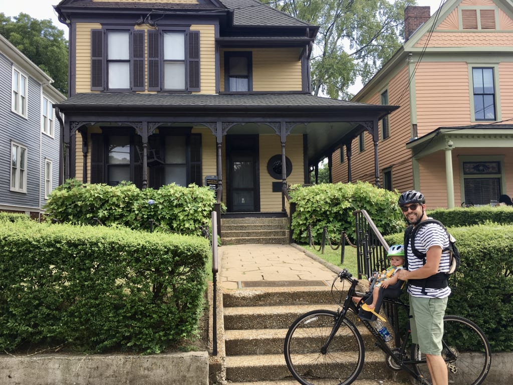 A man on a bicycle with a child in a front seat rides past a Victorian-style house with a porch in Atlanta, Georgia.