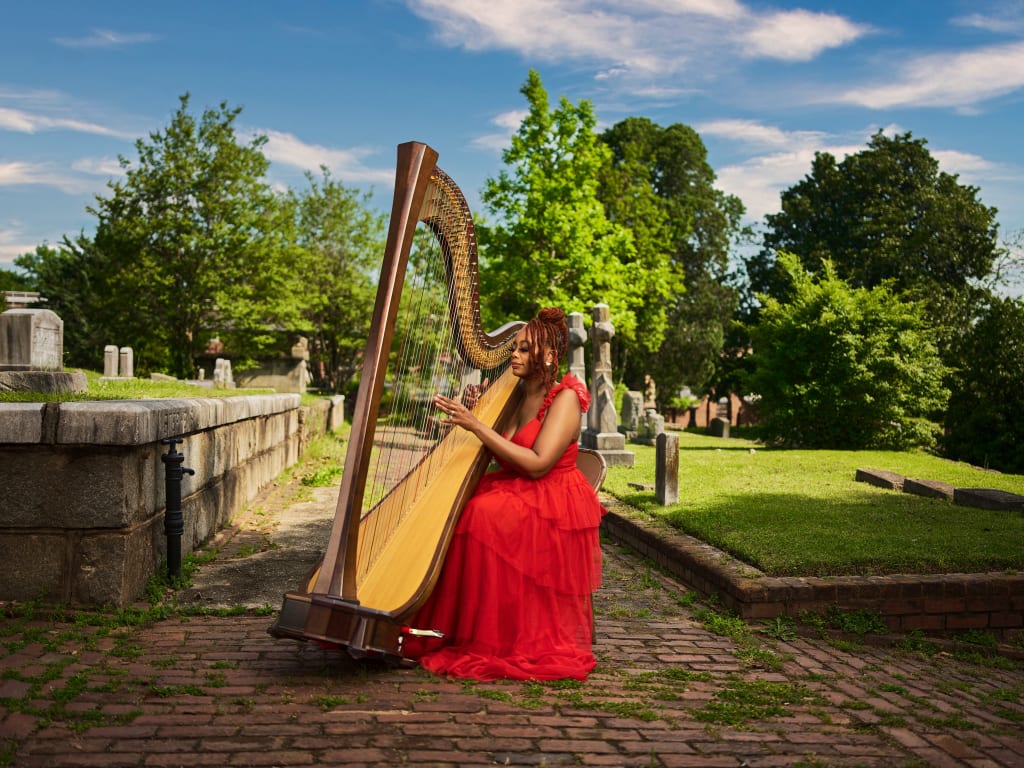 Woman in red dress playing harp in serene cemetery setting with gravestones and trees in Atlanta, Georgia.