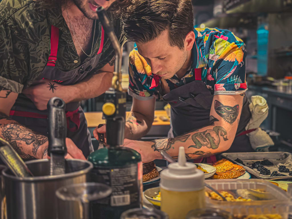 Inside Gunshow Restaurant: How Chef Cody Chassar Leads an Atlanta Icon 6 Two tattooed chefs in aprons prepare food in a busy kitchen with various ingredients and tools in Atlanta, Georgia.
