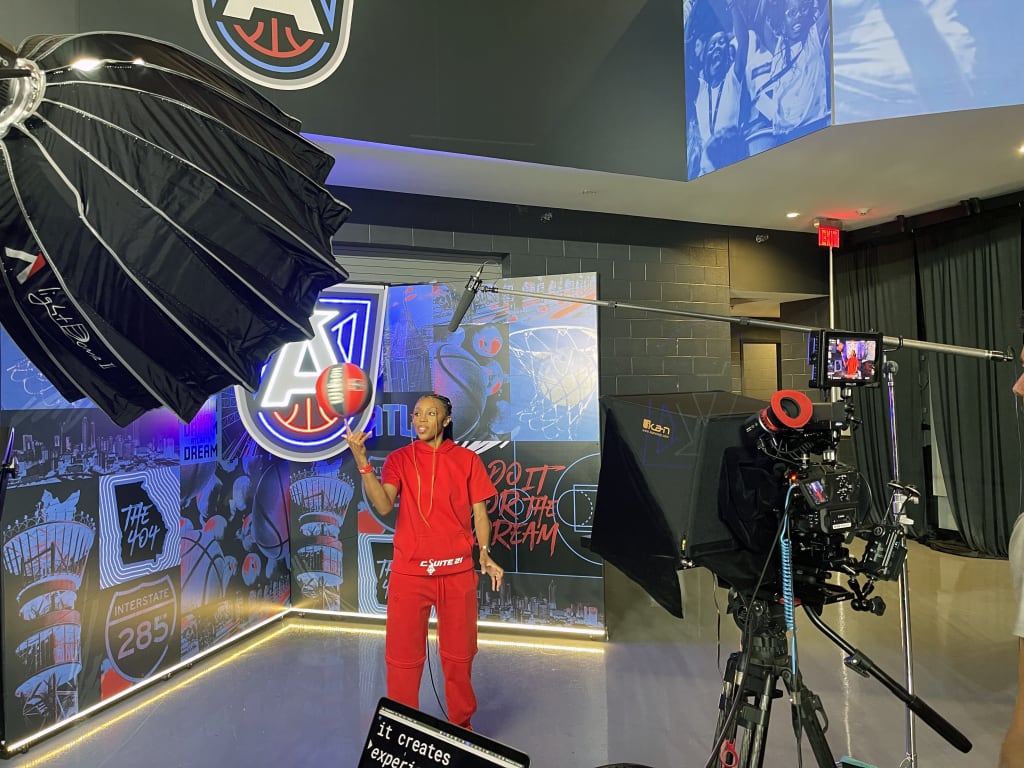 A woman in a red tracksuit poses with a basketball in a studio with a large 'A' logo and cityscape backdrop in Atlanta, Georgia.