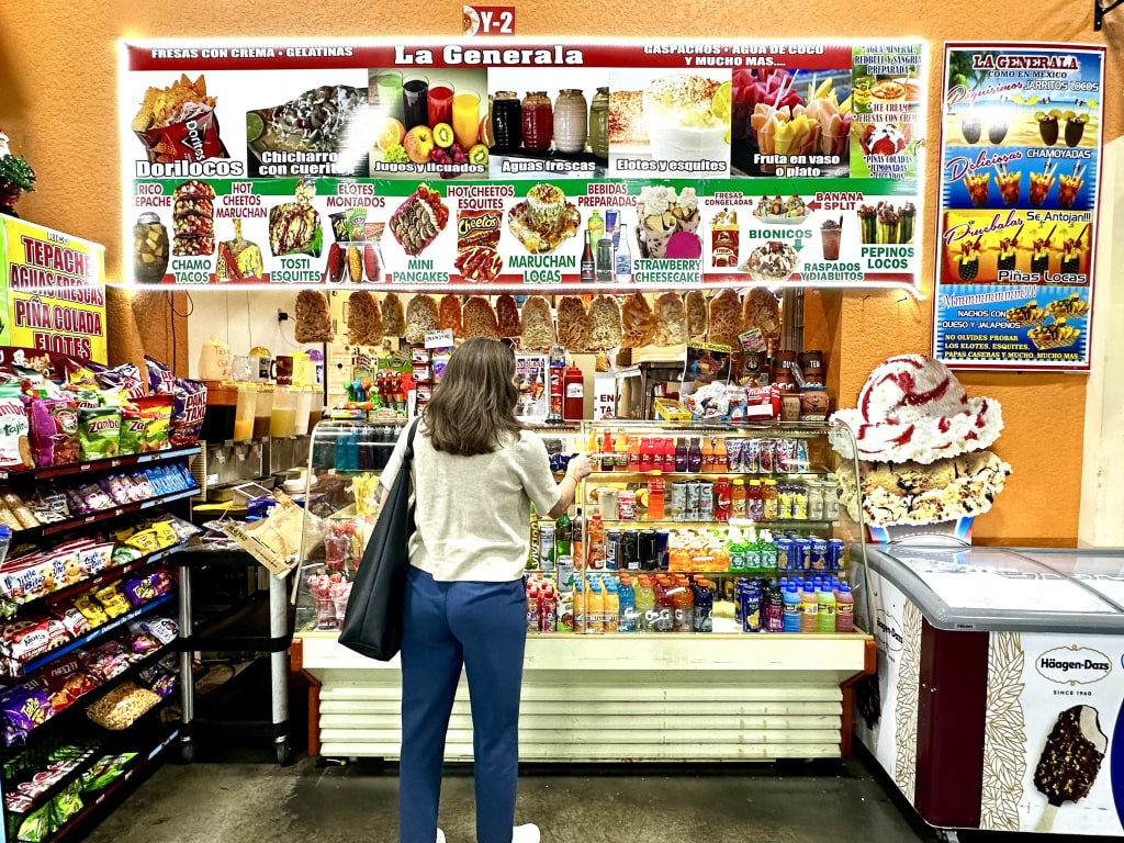 A woman stands in front of a brightly lit, colorful Mexican street food stand named 'La Generalera', which offers a variety of snacks, drinks, and desserts in Atlanta, Georgia.