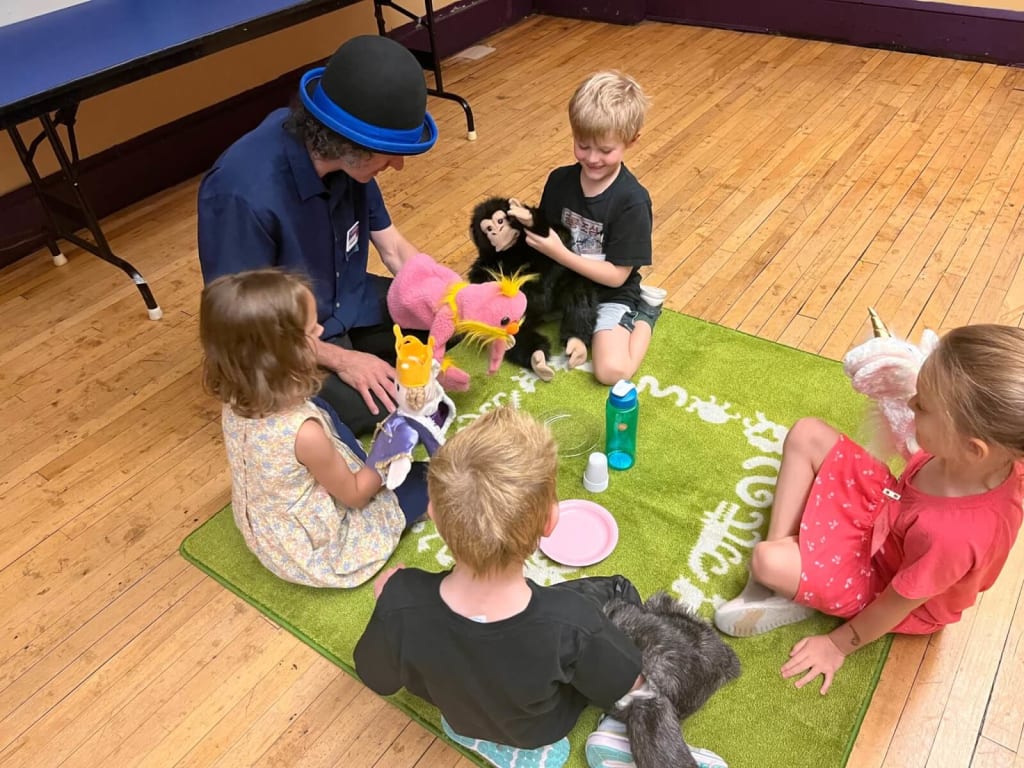 A group of children and an adult are sitting on a green rug, engaged in a playful activity with various stuffed animals and puppets in Atlanta, Georgia.