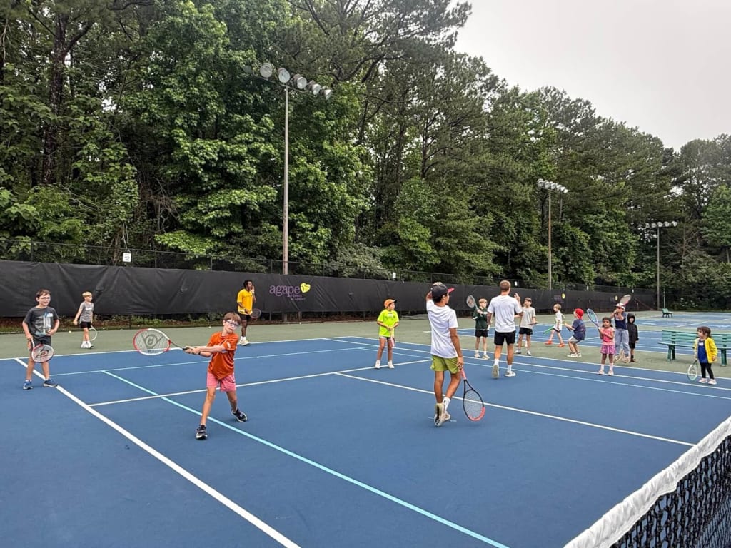 A group of children and adults are playing tennis on a blue outdoor court surrounded by lush green trees in Atlanta, Georgia.