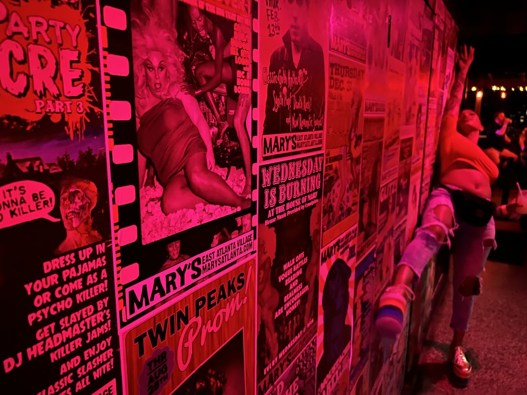 Wall of drag queen show posters and woman posing next to it.