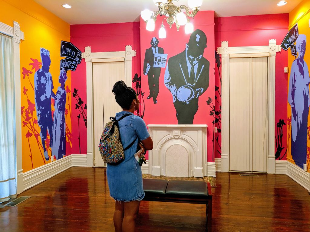 A woman stands in a vibrantly painted room, admiring murals of historical figures and scenes in Atlanta, Georgia.