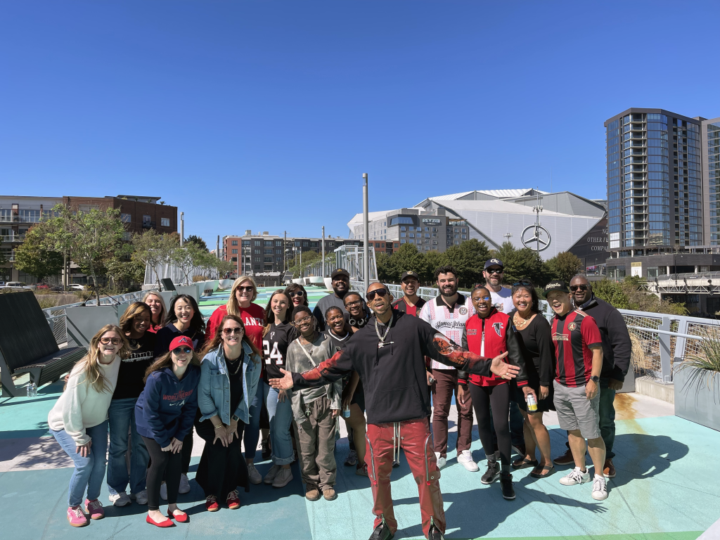 A diverse group of people, dressed in casual and sports attire, pose for a photo on a colorful outdoor platform in Atlanta, Georgia.