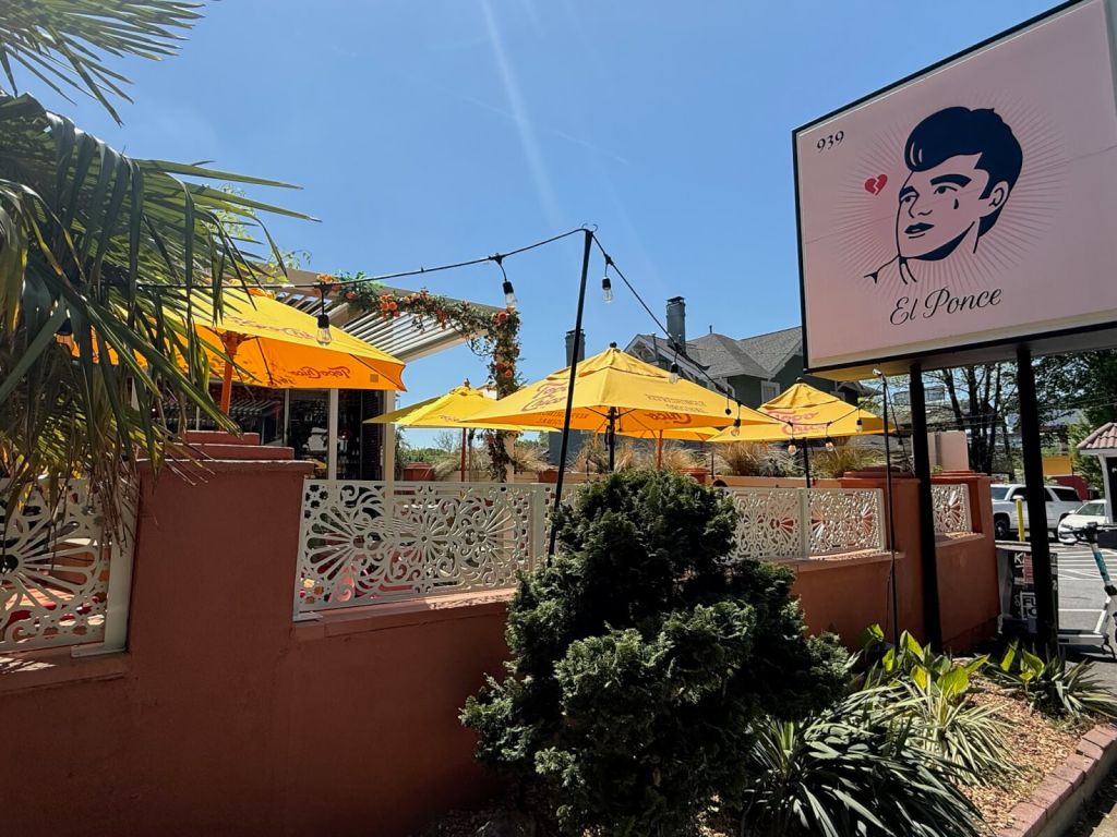 A vibrant outdoor seating area with yellow umbrellas and a sign featuring a stylized man's face with a heart for an eye, set against a backdrop of clear blue skies and lush greenery in Atlanta, Georgia.