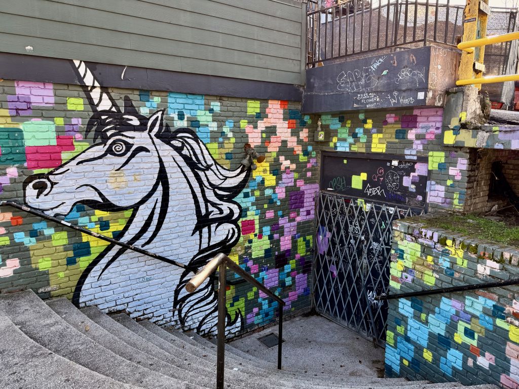 A vibrant mural of a white unicorn with a colorful mane and tail adorns a brick wall next to a staircase, surrounded by a mosaic of multicolored tiles in Atlanta, Georgia.