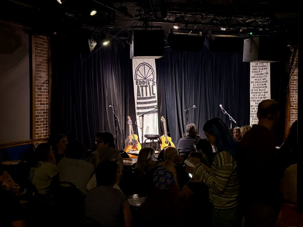 A dimly lit theater stage with a banner reading 'Eddie's Attic' and a crowd of people seated in the audience, ready for a performance in Atlanta, Georgia.