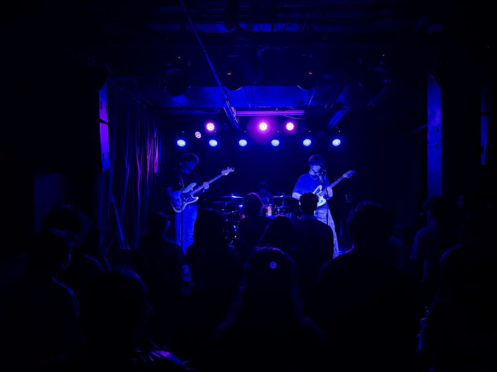 A dimly lit concert venue with a band performing on stage, illuminated by blue and purple stage lights, while an audience watches from the shadows in Atlanta, Georgia.