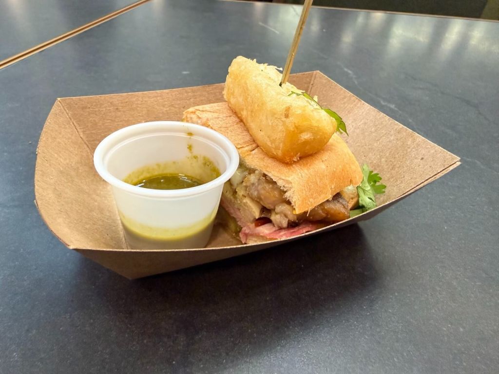 A hearty sandwich with a crispy breaded patty, served with a side of green sauce in a small plastic cup, all presented in a brown paper tray in Atlanta, Georgia.