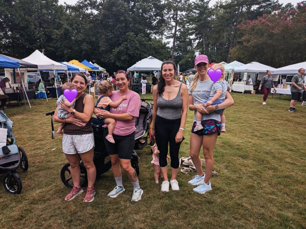 Five women stand together at an outdoor event, each holding a baby or toddler, with colorful tents and a crowd in the background in Atlanta, Georgia.