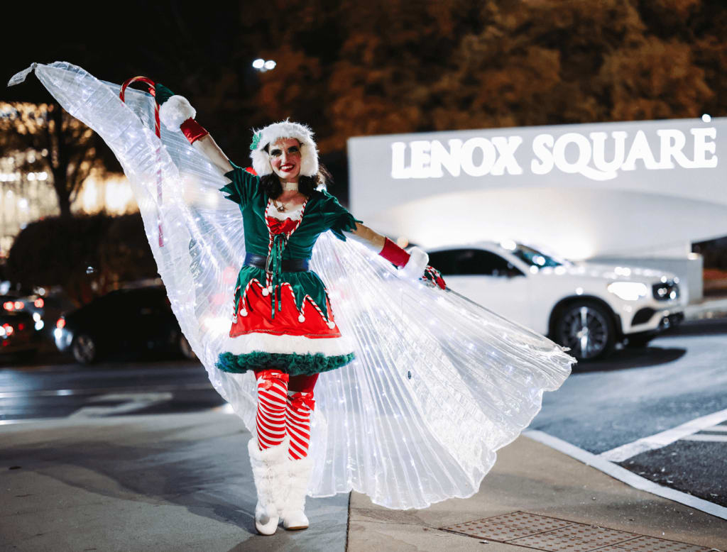 A festive elf performer in a green and red costume with glowing wings dances in front of a car in Atlanta, Georgia.