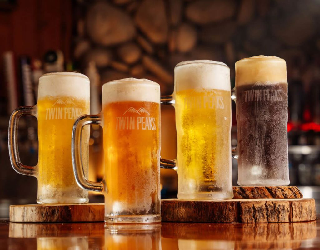 Four frosty beer mugs, each featuring the 'Twin Peaks' logo, are arranged on a wooden bar top, showcasing a variety of beer styles with different colors and head sizes in Atlanta, Georgia.