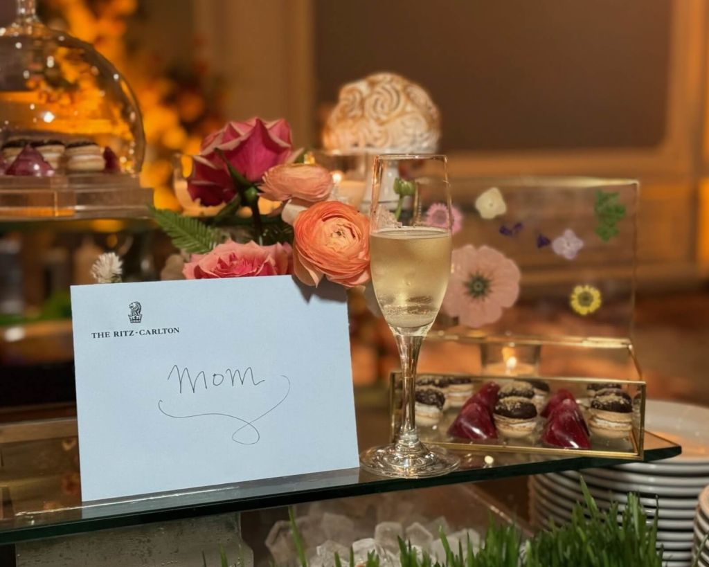 A beautifully arranged table setting at The Ritz-Carlton features a glass of champagne, a handwritten 'Mom' card, and an assortment of colorful flowers and desserts in Atlanta, Georgia.