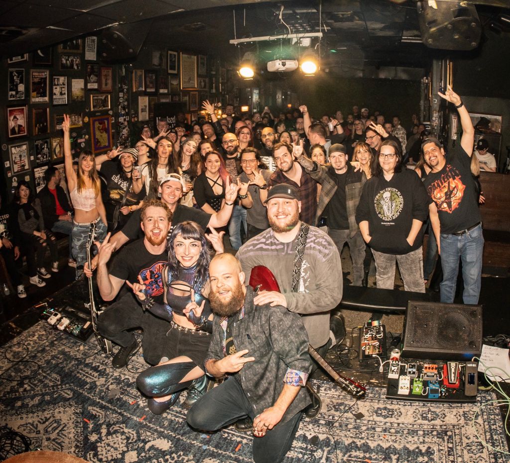 A lively crowd of music enthusiasts, many wearing black and making rock-on gestures, fills a dimly lit venue adorned with posters and framed pictures, creating an energetic atmosphere in Atlanta, Georgia.