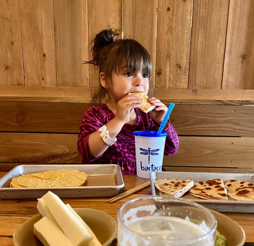 A young girl enjoys a quesadilla with a blue straw at a wooden table in Atlanta, Georgia.
