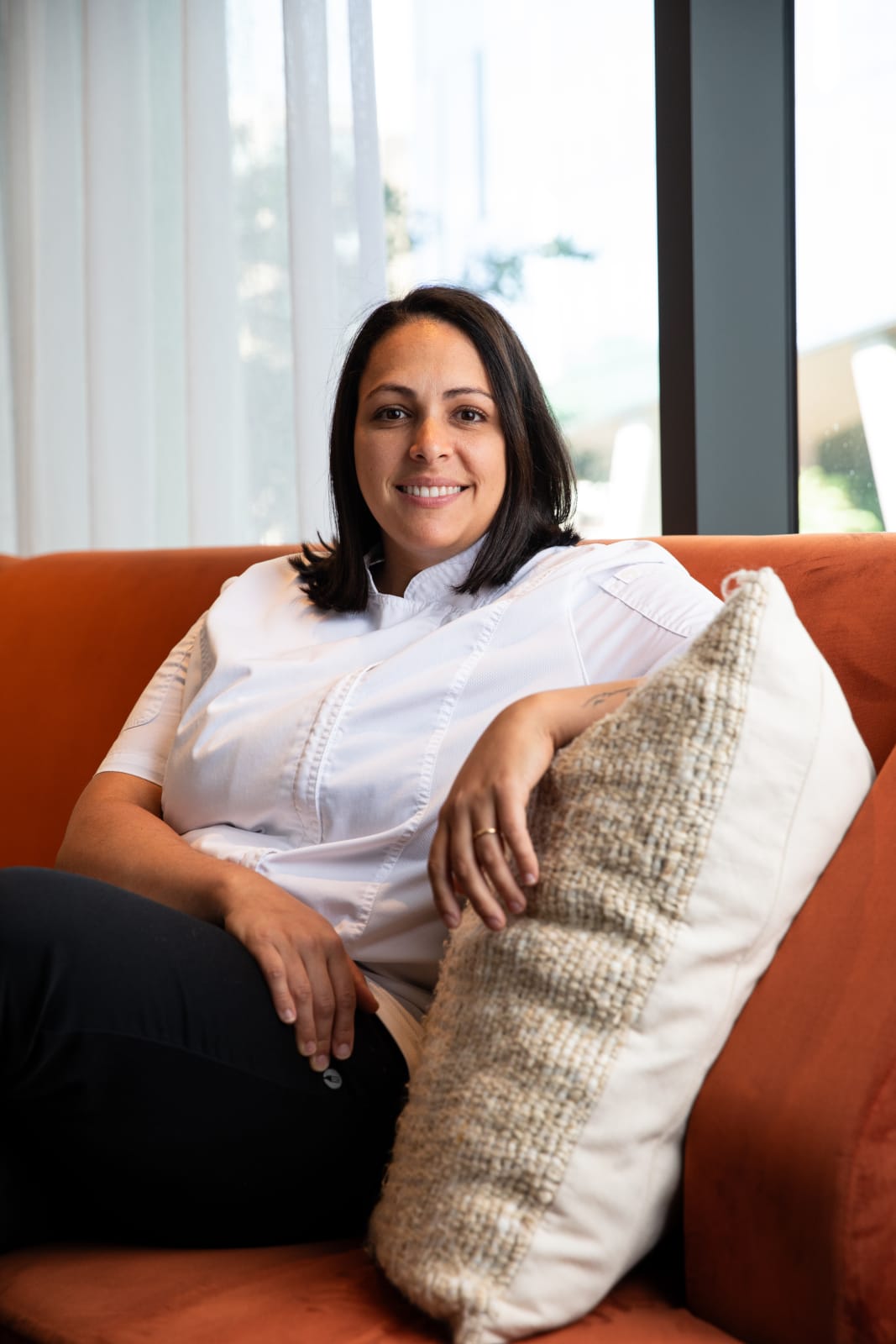 A woman with dark hair smiles while sitting on an orange couch with a white pillow in Atlanta, Georgia.