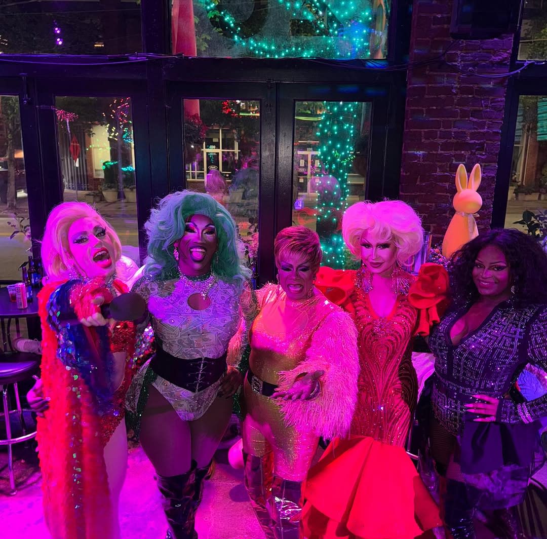 LGBTQ Bars and Nightlife in Atlanta 1 Five drag queens in elaborate costumes pose together in a vibrant, neon-lit bar setting in Atlanta, Georgia.