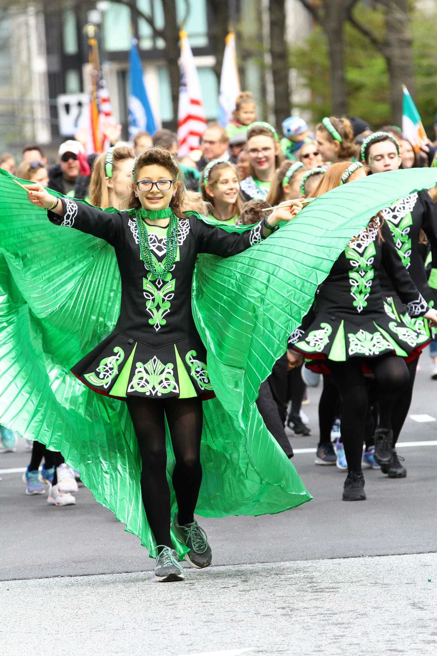 A young woman in a vibrant green and black Irish dance costume leads a parade, arms outstretched in Atlanta, Georgia.