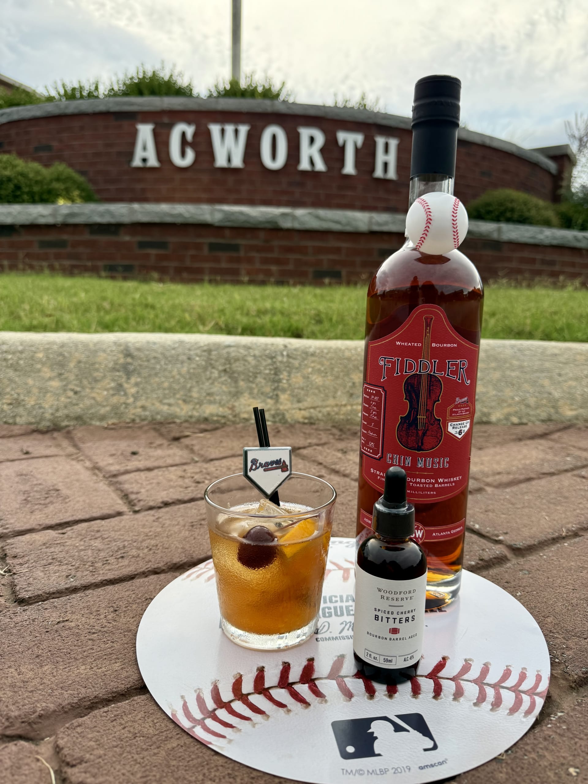 MLB ALL-STAR RESTAURANT WEEK 35 A whiskey cocktail with a cherry and orange slice sits on a baseball-themed coaster next to a bottle of Tiddler whiskey and bitters in Atlanta, Georgia.