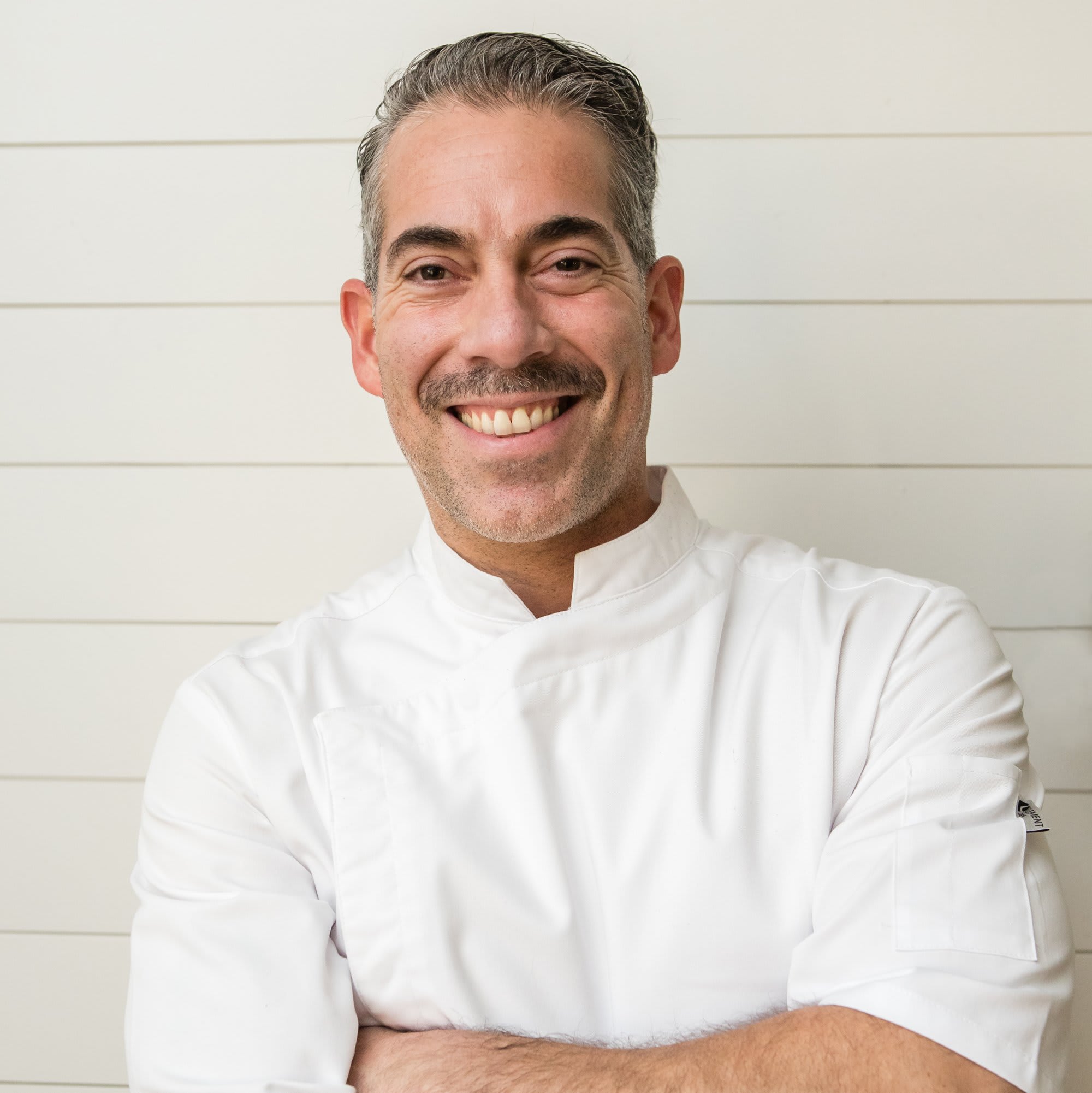 A smiling chef in a white uniform stands with arms crossed against a white wall in Atlanta, Georgia.