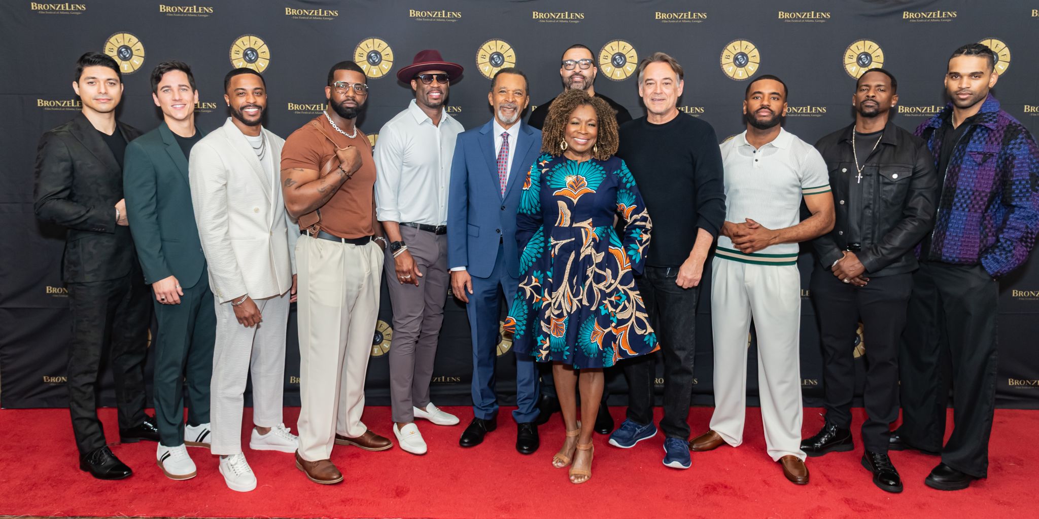 A diverse group of men and women, dressed in stylish formal attire, stand together on a red carpet, smiling and posing for a group photo at a Bronzlens event in Atlanta, Georgia.