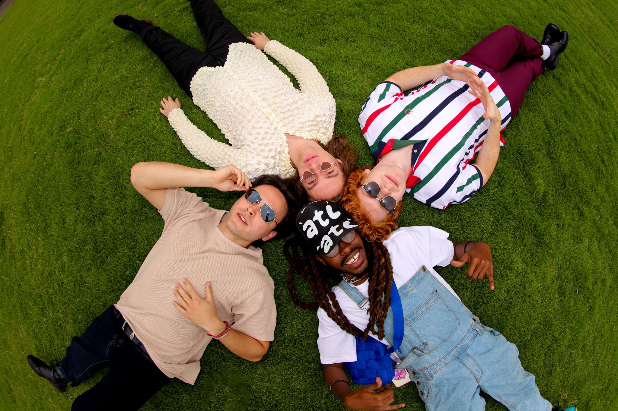 Four friends are lying on their backs on a lush green lawn, each wearing unique and colorful outfits, with sunglasses on, enjoying a sunny day outdoors in Atlanta, Georgia.