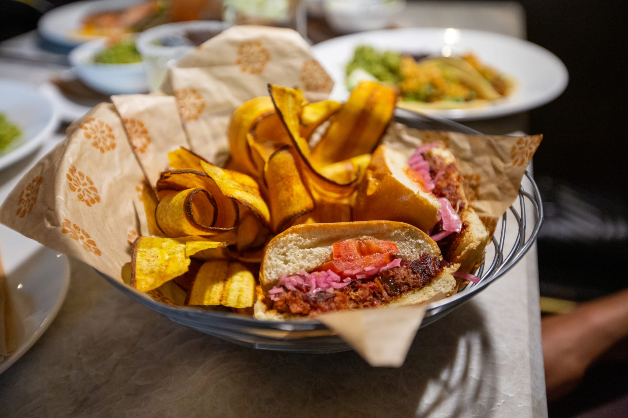 A basket contains a sandwich with meat, tomato, and pickled onions, alongside curly fried plantain chips in Atlanta, Georgia.